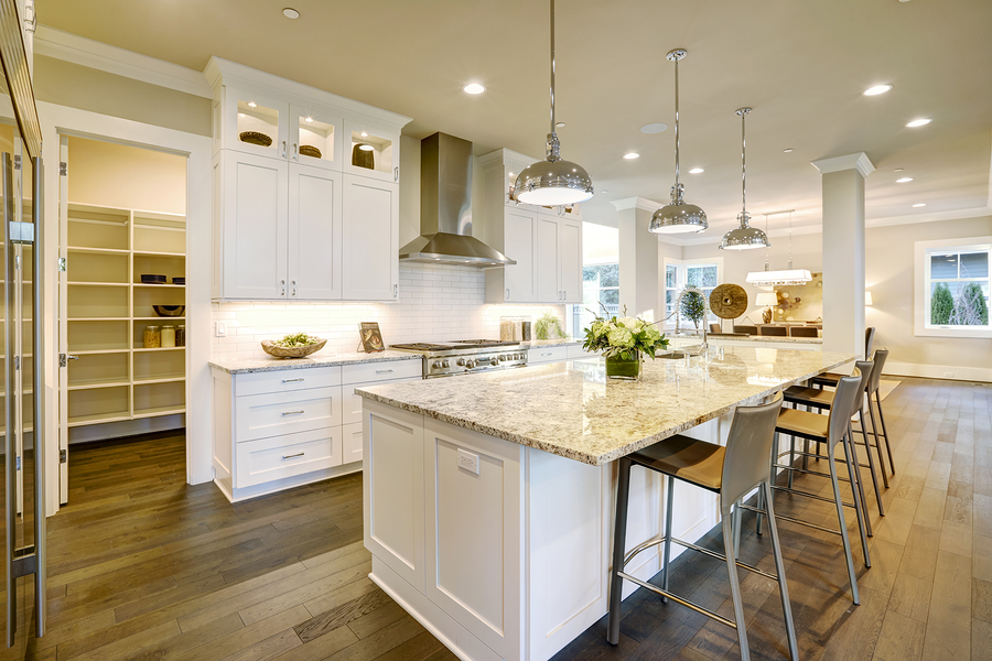 White kitchen design features large bar style kitchen island with granite countertop illuminated by modern pendant lights. Open door lead to walk-in pantry. Northwest USA